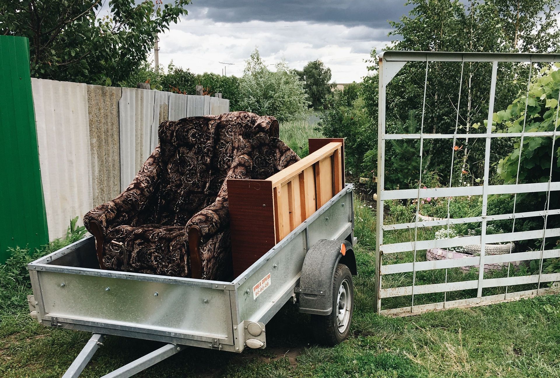 A rural setting with a trailer, vintage armchair, and a pug surrounded by greenery.